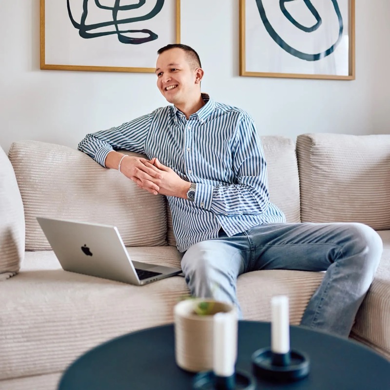 Lukas Zischke lachend auf einem hellen Sofa, daneben ein Laptop